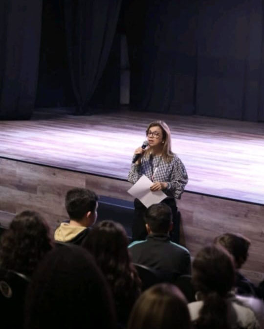 Katia Reis com microfone, falando a um grupo de alunos diante do palco