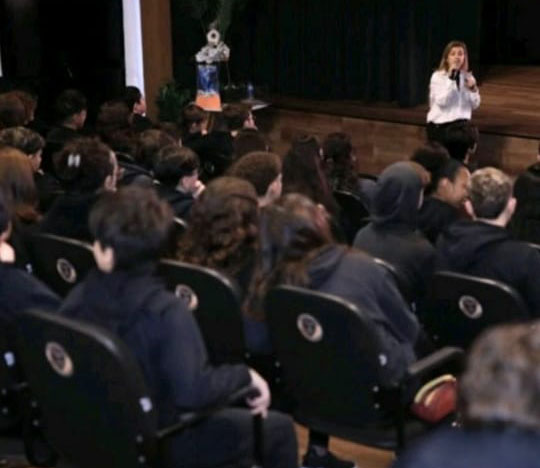 Alunos sentados no auditório assistindo a um encontro conduzido por Katia Reis